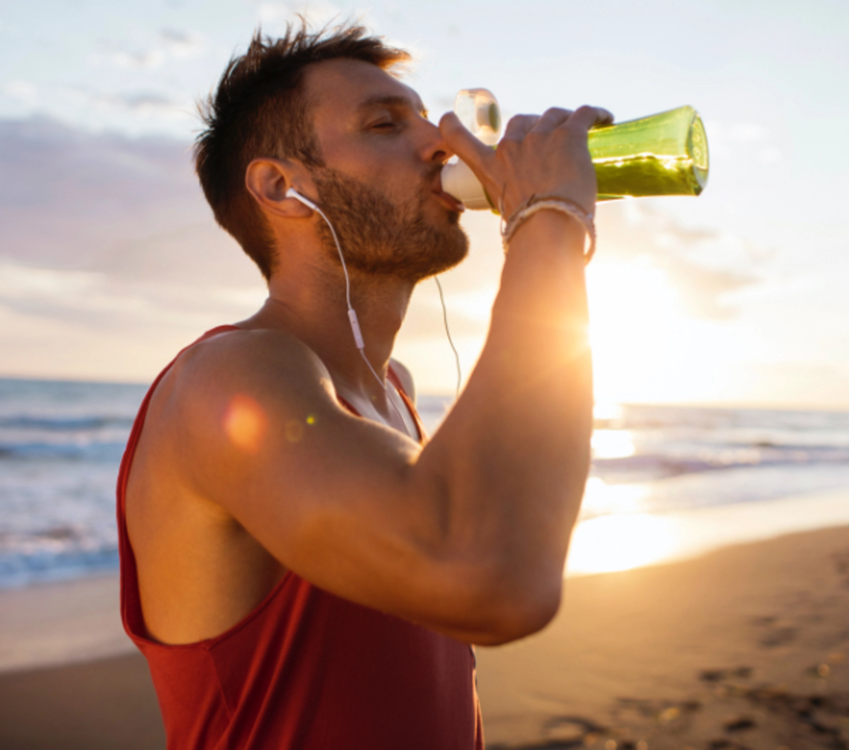 A man in a red exercise shirt drinking out of a green water bottle on the beach.
