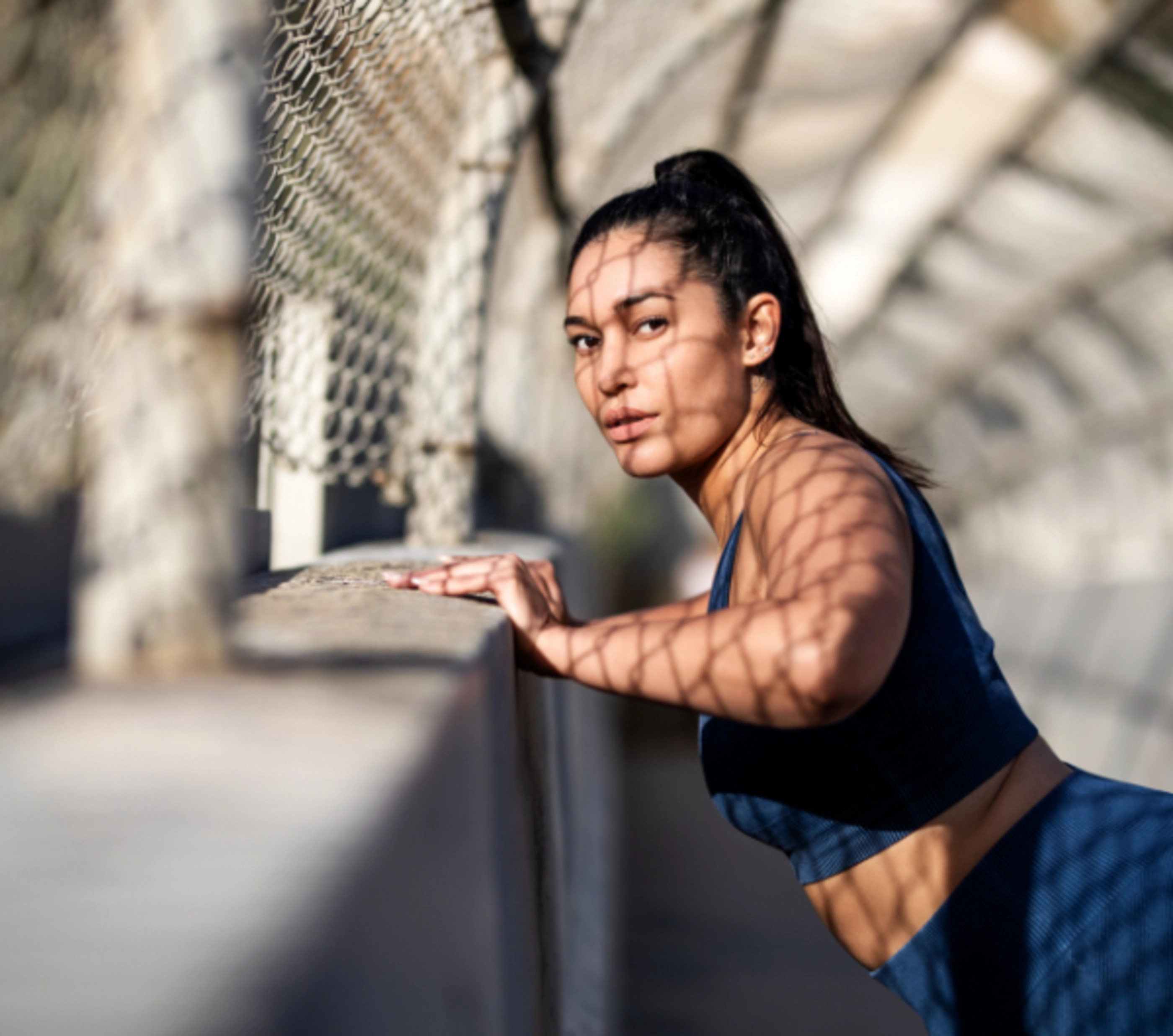 A woman in blue exercise clothes leaning against a cement wall as she stretches.