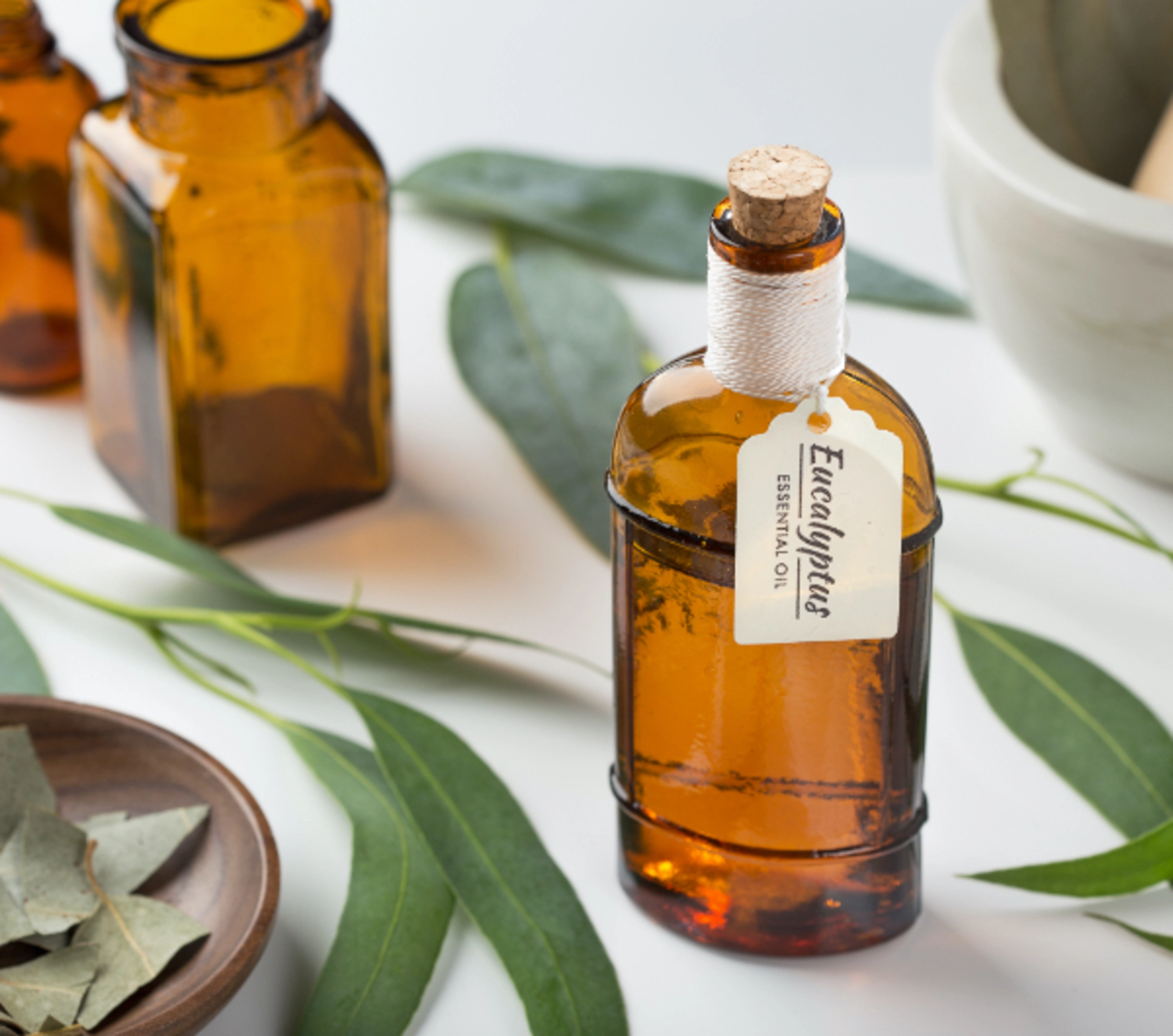 A brown bottle of essential oil with a tag that says 'eucalyptus' sitting on a table covered in eucalyptus leaves.