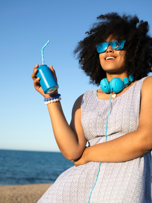 A woman in a blue dress wearing blue headphones and sunglasses drinking out of a blue cup in front of the ocean.