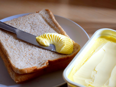A plate with two pieces of bread and a butter knife on it. There is butter on the butter knife and a butter dish next to the plate.