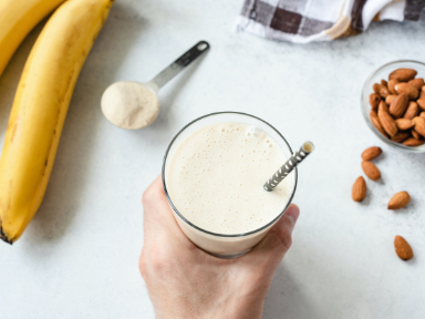 A person holding onto a glass filled with a vanilla milkshake with a bowl of almonds to the right and bananas to the left.