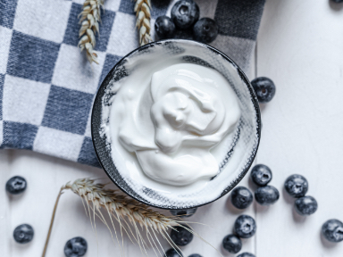 A bowl of yogurt surrounded by blueberries and wheat placed on a blue and white checkered cloth.