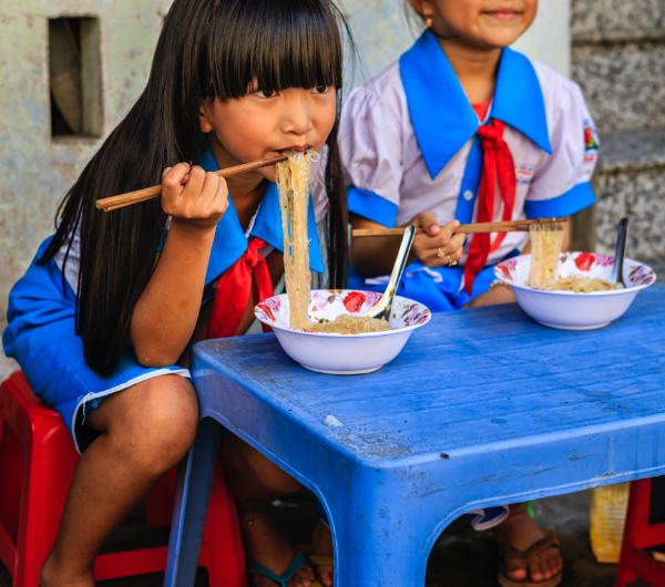 Two young girls in school dresses sitting at a blue table and eating noodles out of bowls with chopsticks.