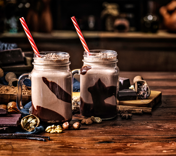 Two chocolate milkshakes in mason jars with red and white striped straws sitting on a wooden table.