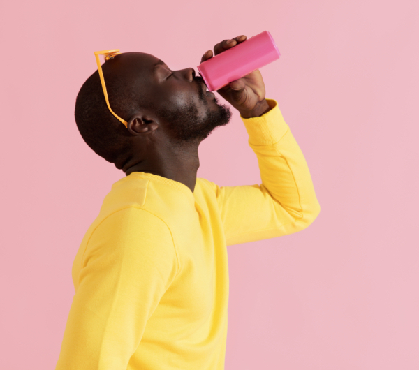 A man wearing a yellow shirt and yellow sunglasses drinking out of a pink glass against a pink background.