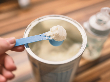 A person scooping infant formula out of a can with a baby bottle in the background.