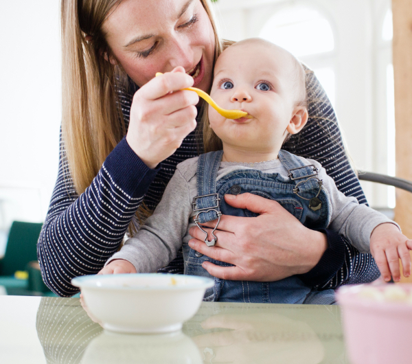 A mom feeding a toddler in overalls baby food.